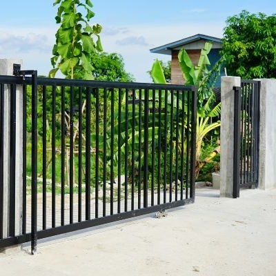 A black metal gate with vertical bars and a latch stands between two grey concrete pillars in sunny Houston. Behind the gate are lush green plants and a small modern structure with a slanted roof. The scene is outdoors under a bright, sunny sky with scattered clouds; perfect weather for any needed gate repair or opener installation.