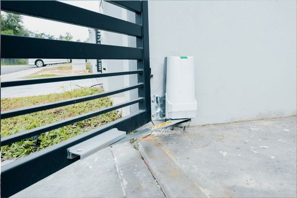 A black metal sliding driveway gate is pictured partially open with a white automatic gate opener device installed on the concrete surface beside it. Green grass and a parked white vehicle are visible in the background, capturing a typical Houston property setup.