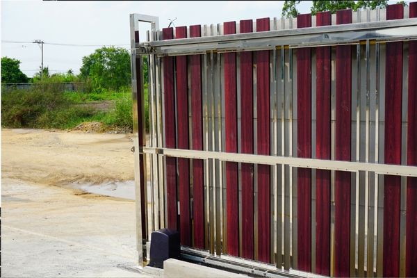 A partially open metal sliding gate, equipped with an electric gate opener, features vertical slats in maroon and silver. The gate sits on a concrete surface, with a rural background including greenery, a dirt path, and a small puddle.