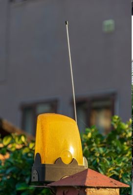 A yellow, rectangular warning light with a small antenna on top, mounted on the corner of a brick wall post near an electric gate. The light is outdoors, with some green foliage and a building with windows in the blurred background.