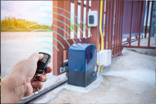 A hand holding a remote control activates an automated sliding gate opener. Blue waves are illustrated to signify the remote signal. The gate is partially open, and the gate control mechanism is visible near the bottom of the gate. Sunlight shines in the background, emphasizing a peaceful Houston morning.