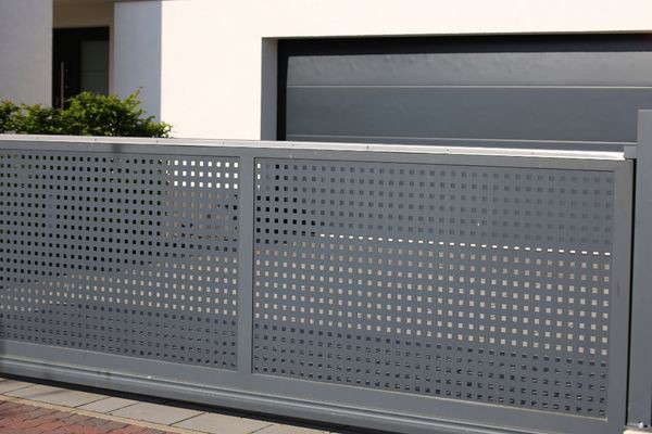 A modern, grey, perforated metal electric gate encloses the driveway of a white building. The gate features a geometric pattern of square holes and is positioned in front of a large, grey garage door. Some greenery is visible in the background, adding a touch of nature to this sleek Houston residence.