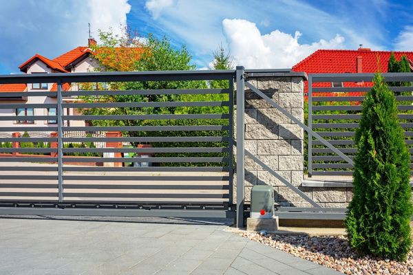 A modern horizontal metal slat electric gate with a stone pillar stands open in front of a residential area. Two houses with red roofs and lush greenery are visible in the background under a partially cloudy blue sky. A small plant is beside the gate.