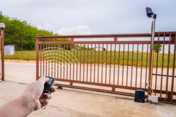 A hand holding a remote control is shown opening an automatic sliding gate with a large, open field in the background. The gate has a simple metal frame design, and a security camera is mounted on a nearby pole. Often tended by gate technicians, the setting appears to be a rural or suburban area.