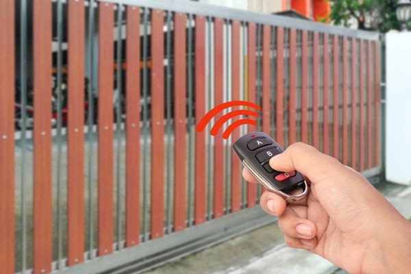 A hand holding a remote control with red signal waves is pointing at a large wooden and metal driveway gate, indicating the gate opener is being used. The gate is partially open, revealing a driveway and some greenery in the background.