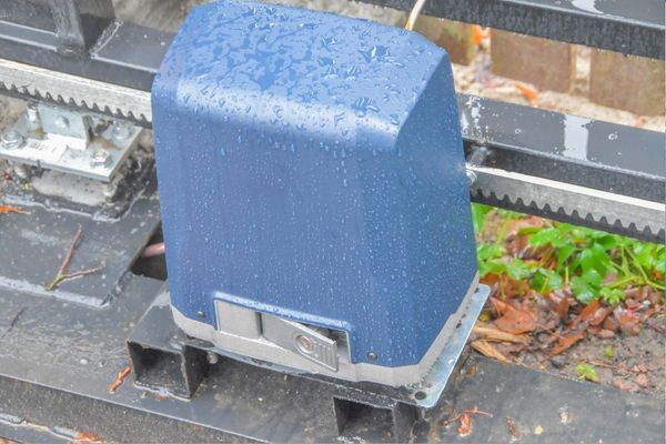 A close-up image of a blue gate motor, part of an electric gate opener system, covered in raindrops. The motor is attached to a metal structure with a toothed rail above it, indicating its role in the automated driveway gate. Wet surfaces and some blurred green foliage can be seen in the background.