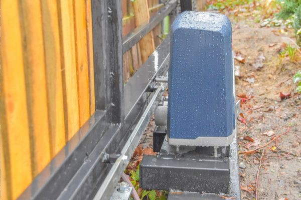 A close-up view of an automatic gate motor installed next to a wooden fence. The motor housing is blue with droplets of rainwater on its surface, and the driveway gate structure is partially visible. The surrounding ground appears wet and is covered with leaves and debris, possibly after a Houston storm.