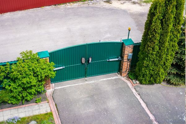 Aerial view of a closed green metal driveway gate set in a brick wall, leading to a paved driveway. The gate is flanked by tall evergreen trees and shrubs, with some greenery visible on the left. A security camera and intercom are mounted on the electric gate.