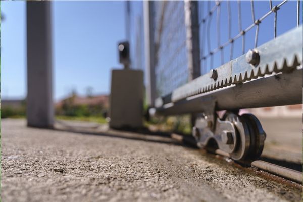 Close-up of the lower part of an electric gate in Houston, showing the gate's wheels on a metal track on a concrete surface. The background is blurred, capturing some greenery and structures under a clear blue sky.