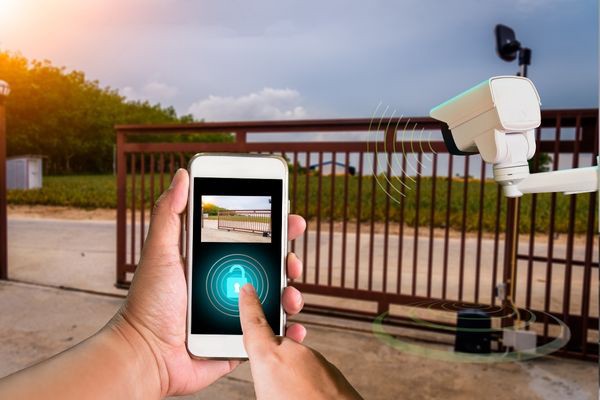 A person uses a smartphone to remotely unlock an electric gate in Houston. The phone screen displays a lock icon and an image of the gate. Nearby, a security camera monitors the area while gate technicians stand by. The background features lush greenery and a clear sky.