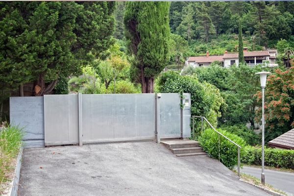 A metal gate equipped with an electric gate opener blocks a driveway that leads to a secluded house surrounded by lush greenery and trees. The gate is flanked by a wall to the left and a hedge to the right. A street lamp stands on the edge of the road that curves down the hillside.