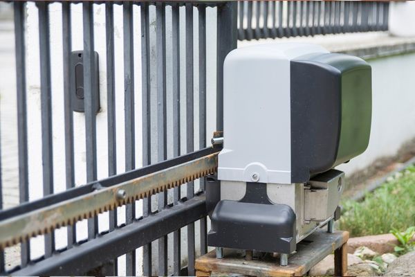 Close-up of a black and grey electric gate opener mechanism attached to a metal gate. The driveway gate features vertical bars and is part of a fenced property. The opener is positioned on a wooden platform near the ground, ensuring efficient operation and ease of potential gate repair services.