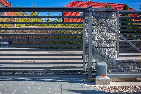 A modern horizontal metal slat sliding gate with an electric gate opener is partly open, revealing a paved driveway and a house with red roof tiles in the background. The gate, spotted in Houston, is mounted on a sturdy stone pillar.