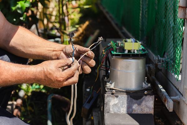 Close-up of a person holding and connecting electrical wires to a motor or electrical device placed on a table outdoors. Nearby, gate technicians work on a driveway gate, with the greenery and green mesh fence in the background, perhaps somewhere in Houston.