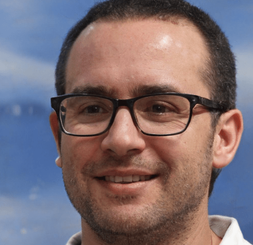 A man with short dark hair and a receding hairline is wearing black-framed glasses and a light-colored shirt. He is smiling, standing by his driveway gate, with the blue Houston sky and some clouds in the background.