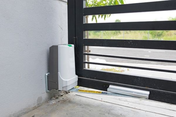 The image shows an automatic sliding driveway gate opener attached to a metal gate on the left side. The mechanism is installed on a concrete surface next to a wall, with a guide rail running along the bottom of the gate. In the background, there is a road and some greenery.
