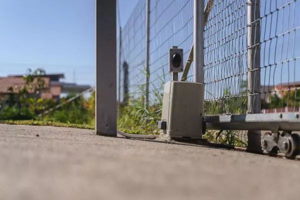 A low angle shot of a metal driveway gate showing its base with a rolling mechanism and gate opener, a security camera mounted nearby, and a background of blurred buildings and greenery under a clear blue Houston sky.