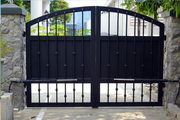 Black wrought iron gate with vertical bars and decorative elements in an arched design, set between two stone pillars. The surrounding area features some greenery and a paved driveway leading up to the gate, which would benefit from the expertise of gate technicians. Buildings and trees are visible in the background.