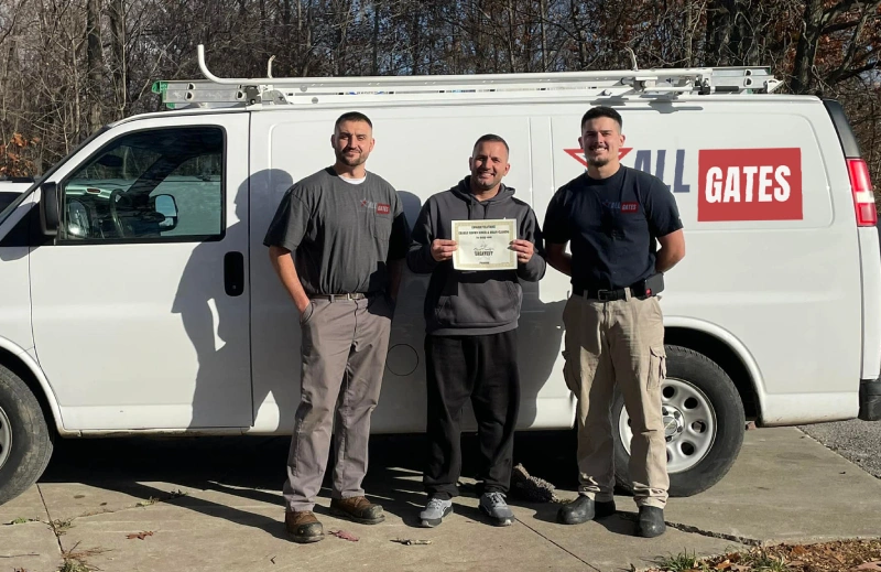 Three men are standing in front of a white work van with the logo “Gates” on it. The man in the center is holding a certificate and smiling. They are wearing work clothes, and two of them have tool belts. The background features trees and parked vehicles.