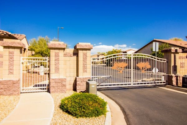 A gated residential community with a wrought iron driveway gate featuring tree designs. The entrance is flanked by stone and brick pillars, and well-maintained shrubbery is visible in the foreground. The road leads into the neighborhood under a clear blue sky.