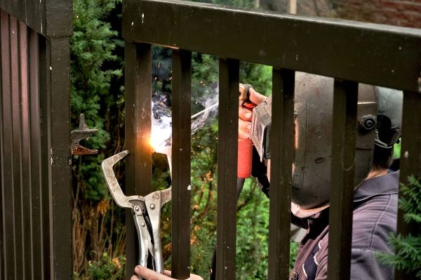 A gate technician, wearing a welding mask, skillfully operates a welding torch and clamp to repair an electric driveway gate outdoors.